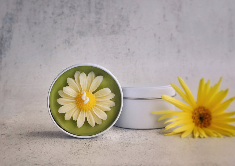 white tin candle, green wax with white petal daisy styled next to another white tin and a yellow flower