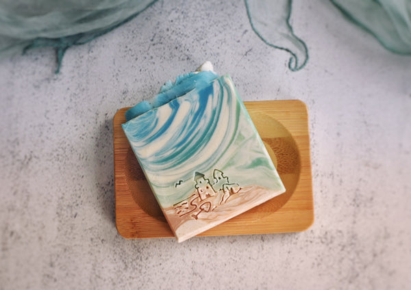 Sea themed soap with sand an blue part with a sand castle stamp on the soap and a soap thong on top of the soap as decoration on a bamboo soap dish 